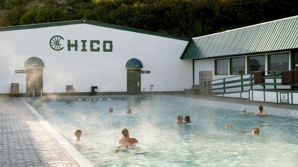 Steam rising off the outdoor hot springs pool at Chico, with the white bathhouse and green roof behind