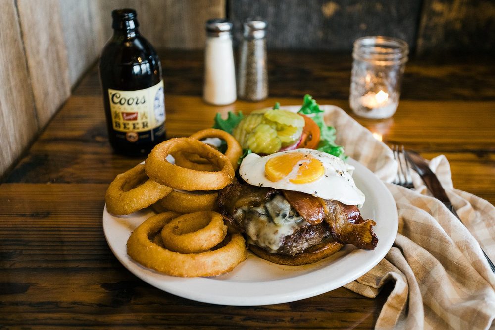 A burger topped with a fried egg and bacon, with onion rings and a Coors bottle on a wood table